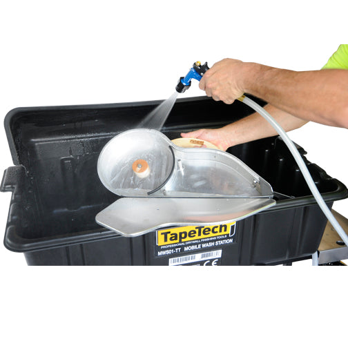 Close-up of a person using a TapeTech water sprayer to clean a taping banjo inside a black wash basin